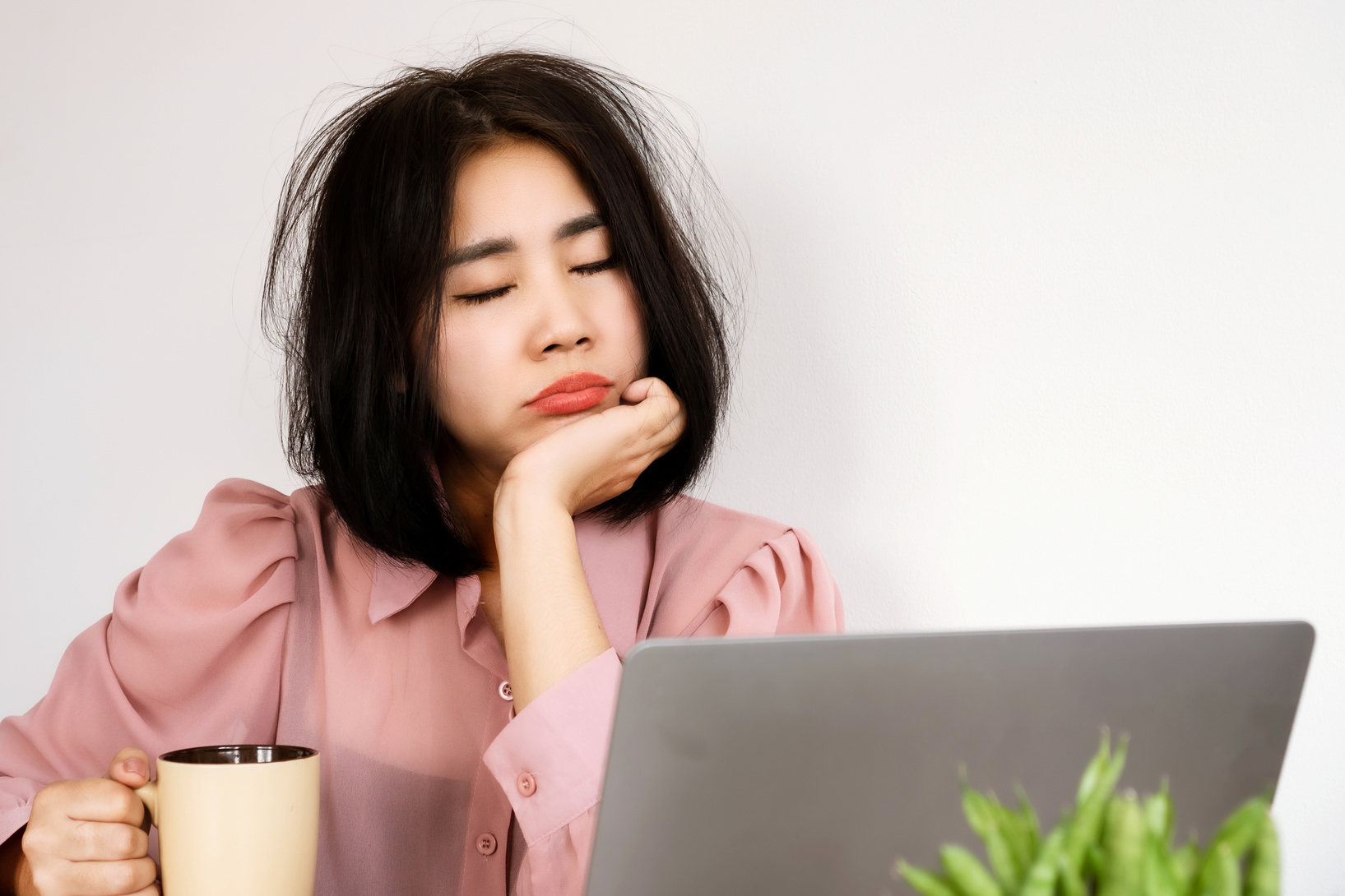 Asian woman Feeling tired or lacking energy after waking up and not feeling refreshed to start the day at workplace hand holding a glass of coffee at office desk